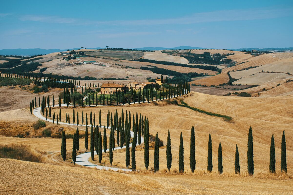 Beautiful Tuscan countryside with a winding, cypress-lined road and rustic villa under a blue sky.